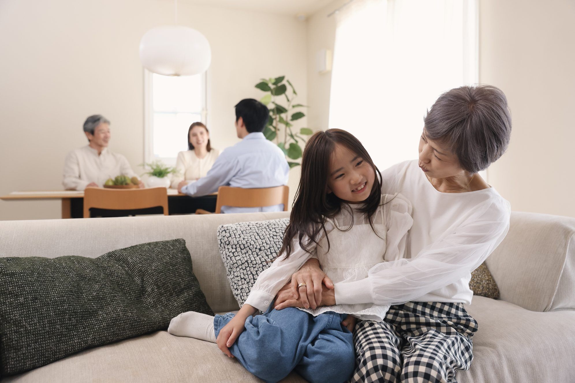 ソファで寄り添う祖母と孫。奥では家族が食卓を囲む